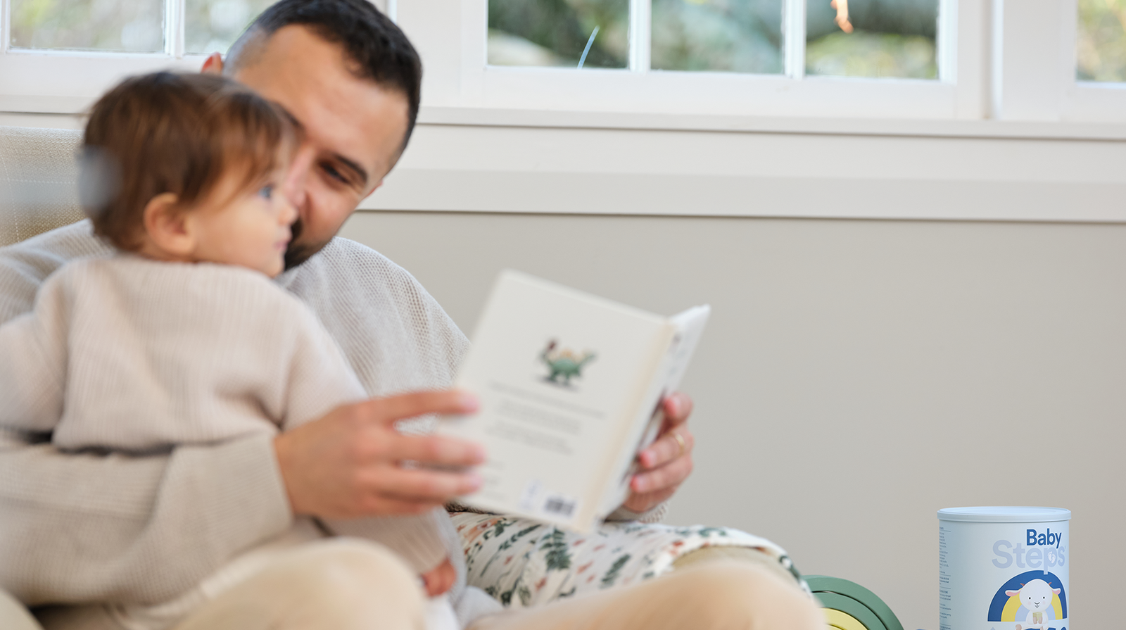 Father reading book to toddler. Baby Steps Stage 3 Milk Drink Can is sitting next to them.