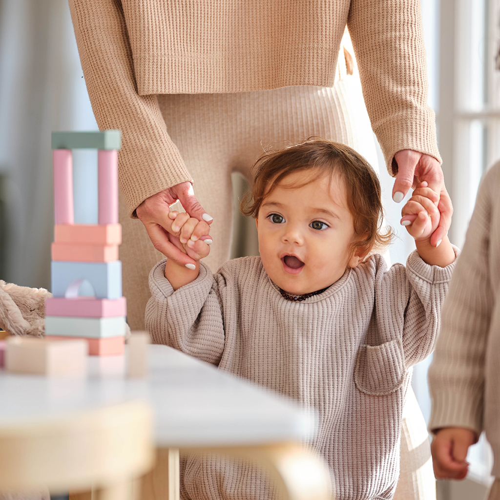 Mother helping toddler walk towards table with wooden toys on. 