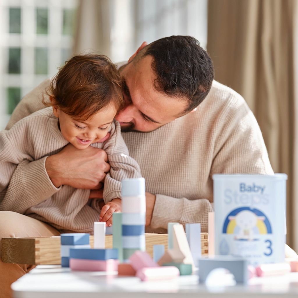 Dad hugging toddler with Baby Steps Stage 3 can sitting on table with wooden toys. 